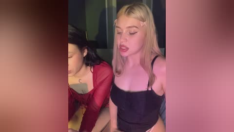 Media: Video of two young women, one with long blonde hair and the other with dark hair, wearing black lingerie and a red top, in a dimly lit room, with pink and red hues.