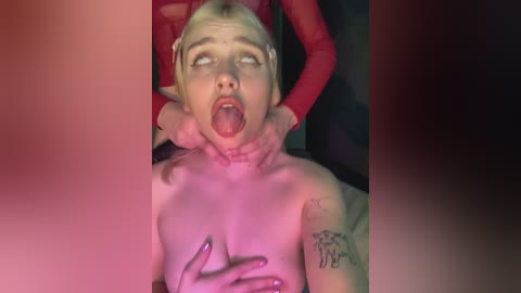 Video of a blonde woman with a small, pierced nose, wearing a red mask, lying on a bed, her breasts exposed, being held down by an unseen person.