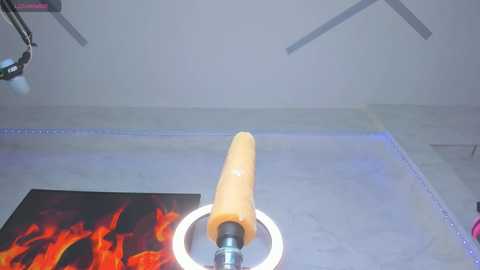 A video of a large, realistic, orange dildo mounted on a metal stand with a glowing blue ring on a gray floor, next to a colorful painting of fire and flames.