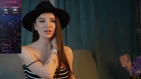 Media: Video of a young woman with long brown hair, wearing a black hat and striped off-shoulder top, standing against a teal curtain background. She appears to be in a cozy, dimly lit room.