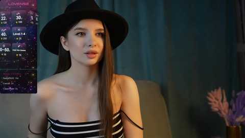 Media: Video of a slender young woman with long straight brown hair, wearing a black hat and black and white striped off-shoulder top, indoors with a dimly lit background.