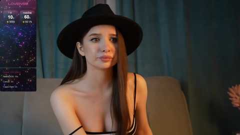 Media: Video of a young woman with long brown hair, wearing a black hat and striped tank top, seated in a dimly lit room with blue curtains.