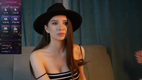 Media: Video of a young woman with long brown hair, wearing a black hat and striped top, indoors by a TV displaying a weather forecast.