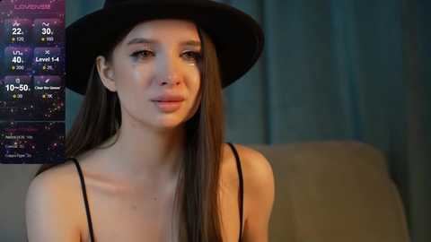 Media: Video of a young woman with long dark hair, wearing a black hat and black top, sitting indoors, with a weather forecast chart in the background.