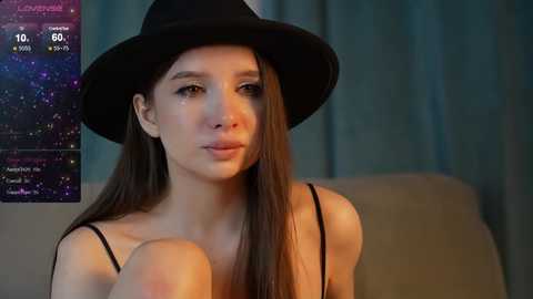 Media: Video of a young woman with long brown hair, wearing a black fedora and black spaghetti-strap top, sitting indoors with a teal curtain background.