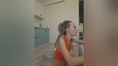 Video of a slender, light-skinned woman with long, blonde braided hair, wearing an orange crop top, kneeling on a gray rug in a minimalist kitchen with white cabinets and a teal sink.