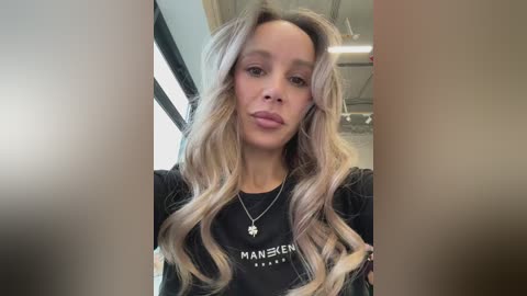 Media: Video of a young woman with wavy, platinum blonde hair, wearing a black \"Mansfield\" t-shirt, standing indoors near a window with sunlight streaming in.