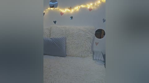 Video of a cozy, white-fur-covered sofa with grey pillows, adorned with heart decals and string lights. Background features a circular mirror and a white wall.