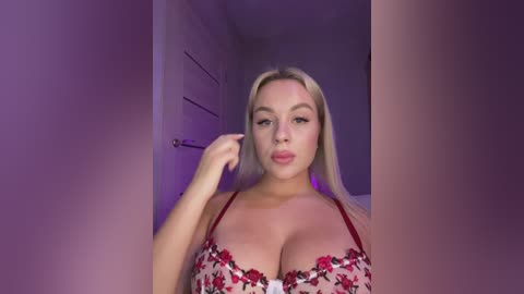 Media: Video of a blonde woman with fair skin, wearing a red floral bra, standing in a dimly lit room with purple lighting. She has a curvaceous figure and full breasts.