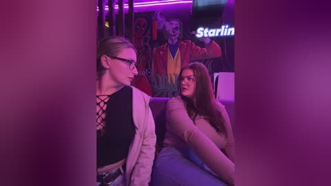 Video of two women in a dimly lit, purple-lit bar. One woman with glasses, black lace-up top, beige jacket, blonde hair, talks to another with long brown hair, beige top, blue jeans, seated on a sofa. Background features colorful mural and \"Starbucks\" sign.
