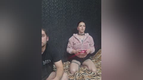 A video shows a young woman with tattoos on her arms, wearing a pink hoodie and black shorts, sitting on a leopard-print blanket, and a man in a black t-shirt, both facing a black, patterned wall.