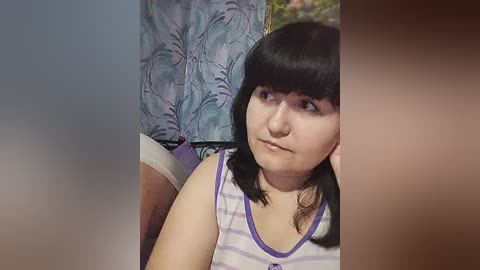 A video of a young woman with straight black hair and fair skin, wearing a white tank top, sitting pensively. The background features a blue and white floral-patterned curtain.