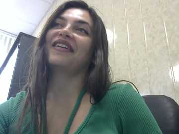 Media: Video of a smiling woman with long brown hair, wearing a green ribbed top, seated indoors against a beige tiled wall with a window in the background.