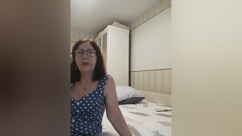 A video of a middle-aged woman with glasses and shoulder-length dark hair, wearing a blue polka-dot dress, sitting on a bed with white and beige patterned sheets in a minimalist room with yellow and white striped wallpaper.