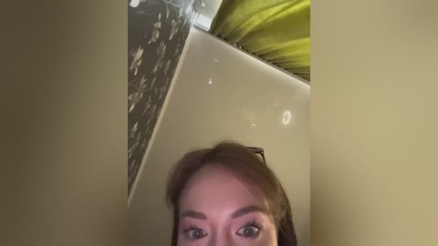 Media: Video of a close-up, slightly blurry face with light brown hair, peering through a gap between a beige wall and a silver metallic surface.