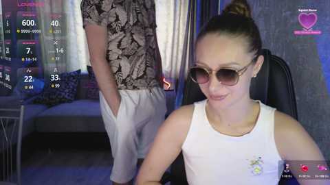 Video of a white woman with brown hair in a ponytail, wearing sunglasses and a white tank top, sitting in a black chair in a dimly lit room with a man in a patterned shirt and white pants standing behind her.