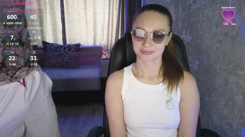 Video of a young, light-skinned woman with a ponytail, wearing sunglasses and a white tank top, sitting on a black chair in a dimly-lit room with a bed and curtains in the background.