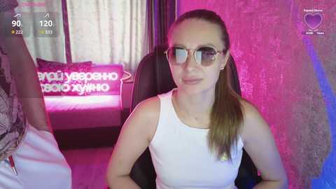 Video of a fair-skinned woman with long brown hair in a ponytail, wearing oversized glasses and a white tank top, seated on a black leather chair. Pink and purple neon lights illuminate the room.