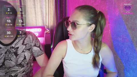 Video of a young Caucasian woman with light skin and long brown hair in a ponytail, wearing sunglasses and a white tank top, sitting next to a person in a patterned shirt, in a room with pink and purple lighting.