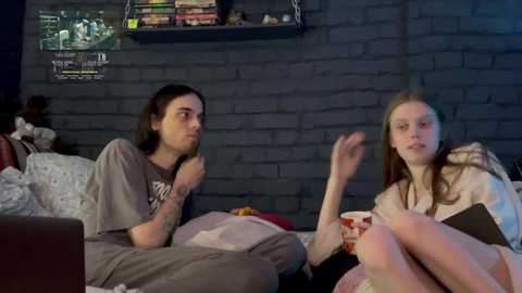 Media: Video of two young people, a man with long hair in a gray t-shirt and a woman with long blonde hair in a beige top, sitting on a bed in a dimly-lit room with a brick wall, shelves, and a \"Doctor Who\" poster.