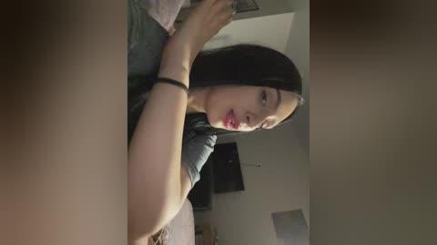 Media: A video of an East Asian woman with long black hair, fair skin, and red lipstick, wearing a grey T-shirt, lying on a bed in a dimly lit room with a TV and posters on the wall.