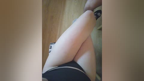 Media: Video of a woman's legs in black shorts, sitting on a wooden floor, with a blurred background.
