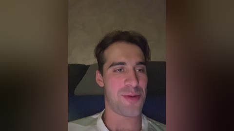 Video of a young Caucasian man with short dark hair and a trimmed mustache, wearing a white shirt, seated indoors on a dark couch against a beige wall.