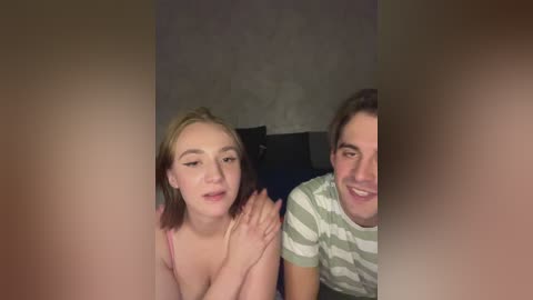 Video of a young Caucasian woman with blonde hair and a man with brown hair, both smiling. They are sitting on a bed, with a dark blue pillow visible. The background is dimly lit with a textured gray wall.