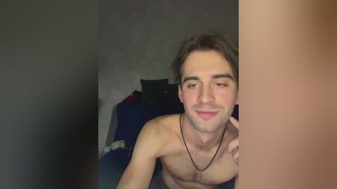 Video of a shirtless young man with light skin and short brown hair, smiling slightly, sitting on a bed with dark blue sheets and pillows, wearing a black necklace.