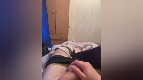 A video shows a man lying on a bed, partially undressed, with a hand on his crotch, surrounded by a beige room with wooden furniture and a blue light source.