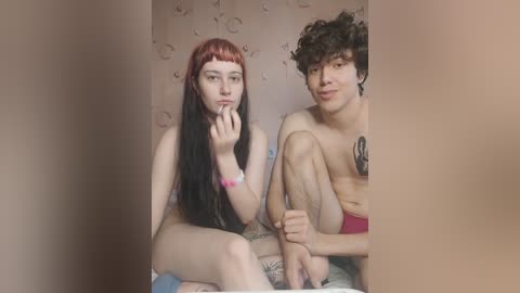 Media: Video of a fair-skinned woman with long red hair, sitting nude next to a shirtless man with curly hair and a tattoo, both smiling, in a room with floral wallpaper.