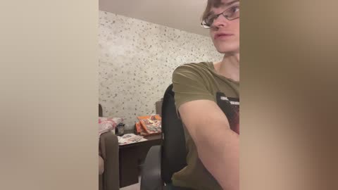Video of a young, fair-skinned man with glasses, short brown hair, wearing a green t-shirt, seated at a desk in a cluttered, wallpapered room.