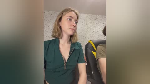 Video of a young, fair-skinned woman with shoulder-length blonde hair, wearing a green zipped-up dress, standing indoors near a black and yellow chair, with a speckled wallpaper background.