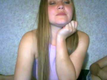 Video of a young Caucasian woman with long blonde hair, wearing a light purple tank top, leaning forward with her chin resting on her hand, against a textured, light-colored wall.
