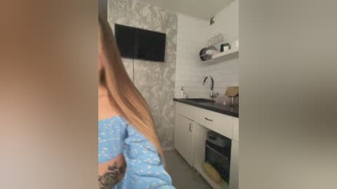 Video of a woman in blue pajamas standing in a modern, minimalist kitchen with white tiles, black countertops, and a black TV mounted on the wall.