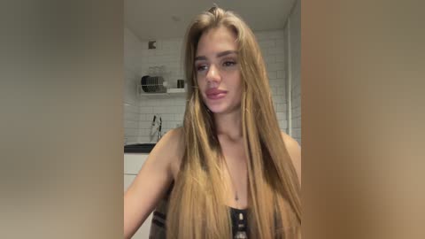 Video of a young woman with long, straight, blonde hair, wearing a strapless top, in a modern kitchen with white tiled walls and stainless steel appliances.