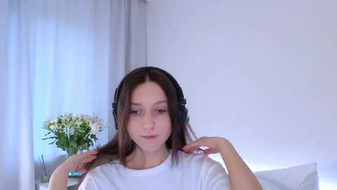 A video of a young woman with fair skin, long brown hair, wearing black headphones, a white T-shirt, and a neutral expression, seated indoors with a vase of white flowers on a white surface.