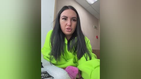 A video of a young woman with long black hair and fair skin, wearing a bright green hoodie and pink and white knitted gloves, standing in a hallway with beige walls and a ceiling light.