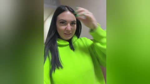 Video of a young woman with long black hair, wearing a bright neon green hoodie, adjusting her hair, standing in a blurred, green-toned room.
