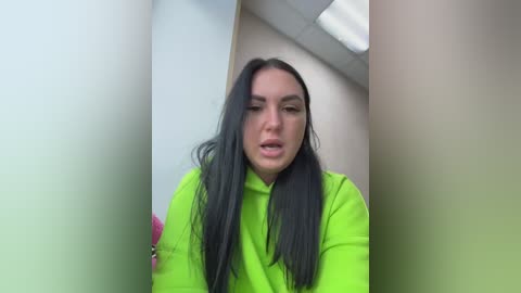 Video of a woman with long, straight black hair, wearing a bright neon green hoodie, standing in a narrow, beige-walled hallway with fluorescent lighting overhead.