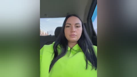 Video of a woman with long black hair, wearing a bright neon green jacket, sitting in a car, with a blurred cityscape visible through the window.