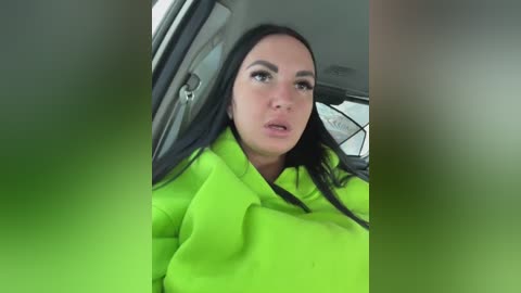Video of a woman with long black hair, wearing a neon green jacket, sitting in a car with blurred green-tinted window frames, looking slightly startled.