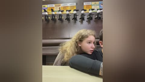 Media: Video of a blonde woman with curly hair, wearing a black sweater, resting her head on a man's shoulder in a dimly lit, institutional setting with wooden walls and a row of office chairs.