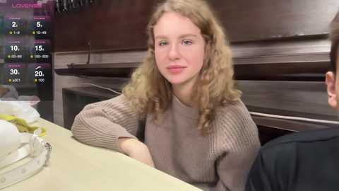 Media: Video of a blonde woman in a brown sweater, smiling, seated at a table with a black person partially visible. Background shows a dark room with a window.
