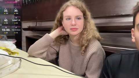 Media: Video of a young blonde woman with curly hair, wearing a beige sweater, seated at a table with a man. Background features a dark wooden wall and a digital screen showing weather and location information.