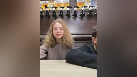 Media: Video of a blonde girl with curly hair, wearing a gray sweater, sitting at a table in a radio studio. Black microphones hang from the ceiling.