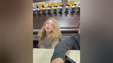 Media: Video of a blonde girl with curly hair in a grey sweater, seated at a wooden table in a classroom with a teacher visible behind her.