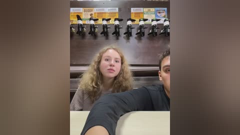 Media: Video of a blonde woman with curly hair in a gray sweater sitting at a table, with a man in a dark shirt visible in the background. A row of black and white soda dispensers and signs are mounted on a dark wall behind them.