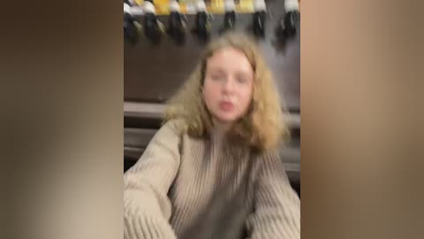 Media: A blurred video of a fair-skinned woman with wavy, shoulder-length blonde hair, wearing a beige ribbed sweater, sitting in a dimly lit room with a black-and-yellow patterned backdrop.