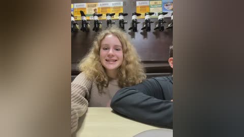 Media: Video of a young woman with curly blonde hair, smiling, wearing a beige sweater, seated at a table in a bar with multiple beer taps in the background.